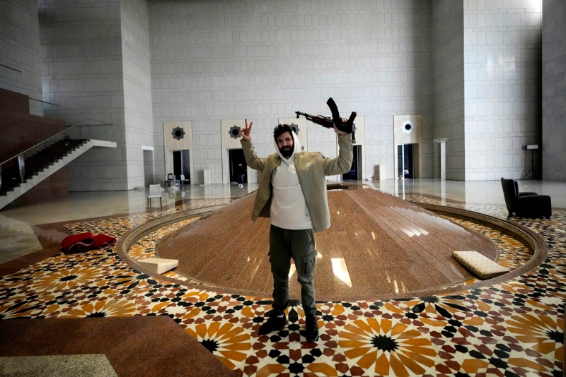 敘利亞總統(tǒng)巴沙爾·阿薩德（Bashar al-Assad）搭機(jī)逃亡后，敘利亞叛軍及民眾闖進(jìn)總統(tǒng)府洗劫，還發(fā)現(xiàn)阿塞德有一整排賓士、法拉利、保時(shí)捷等豪車收藏。圖為8日一名男子手持AK-47在總統(tǒng)府拍照。